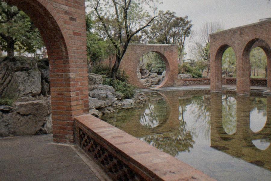 A long corridor of red brick with circular openings leading toward a garden pond